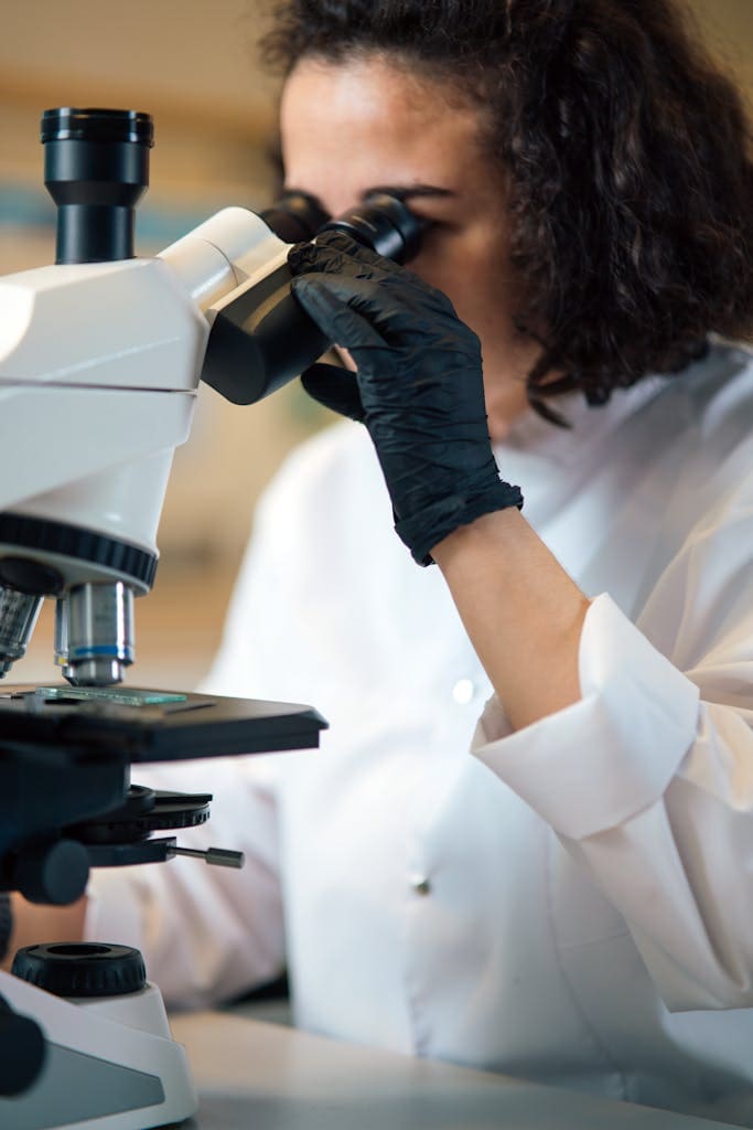 A female scientist examines samples using a microscope wearing gloves in a modern lab.