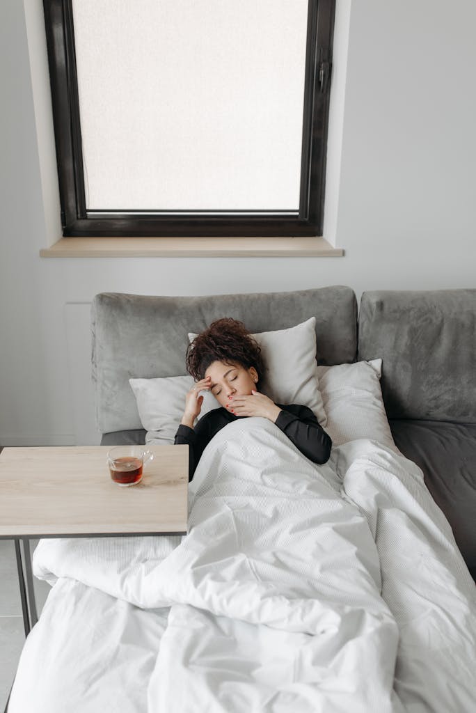 A woman rests in bed, appearing unwell, with a cup of tea nearby for comfort.