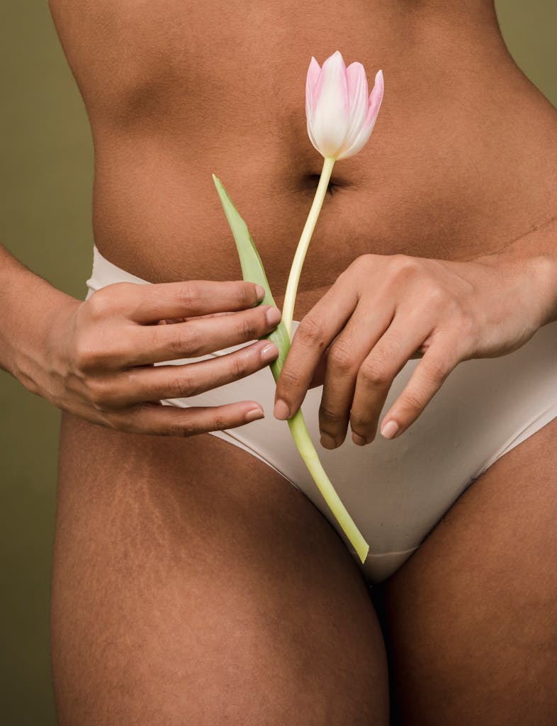 Crop faceless female in white panties with stretch marks on hips holding aromatic tulip near belly against beige background