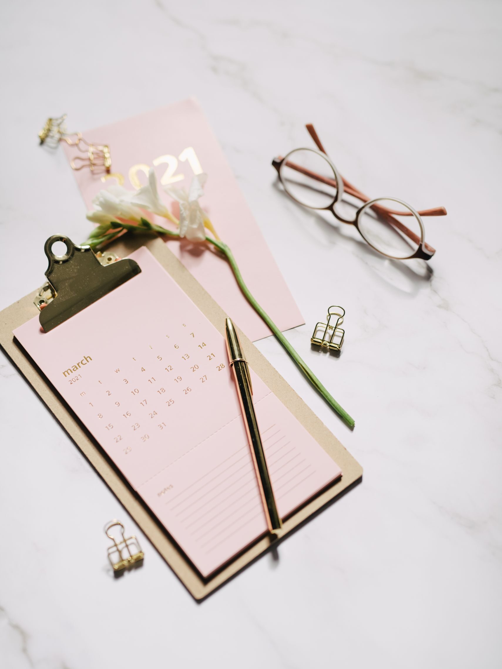 Elegant stationery arrangement on marble surface with calendar, pen, glasses, and flower.
